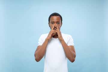 Fototapeta na wymiar Half-length close up portrait of young african-american male model in white shirt on blue background. Human emotions, facial expression, ad concept. Covering face with his hands, looks serious.