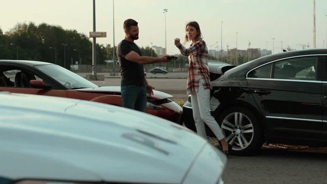 Man and woman drivers talk with anger argue on the car accident look at each other point on the broken vehicles stand on the roadside. Police on background. Road accident.