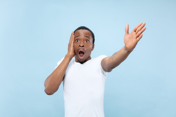 Half-length close up portrait of young african-american man in white shirt on blue background. Human emotions, facial expression, ad, sales concept. Pointing, choosing, astonished. Copyspace.