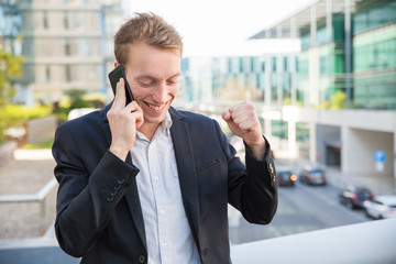 Excited happy business leader celebrating success while talking on phone. Joyful amazed young man...
