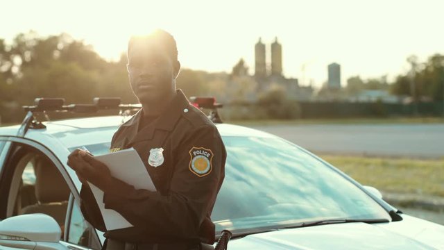 Serious black policeman writing notes working outdoors leaning on the police car hood posing at sunlight looking on camera in the city street.