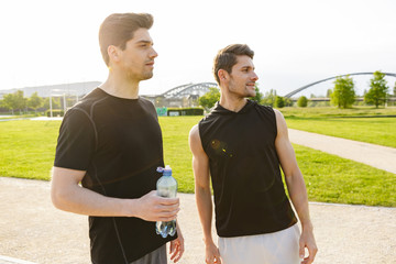 Image of two muscular guys drinking water while working out and running at at green park outdoors in morning