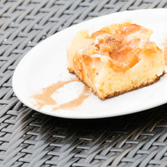Tasty sheet cake with apricots on a white tableware. On the plate there is a cinnamon mask of a spoon visible. Around the white plate there is the dark grey rattan table.  