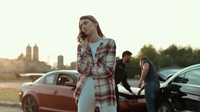 Portrait of impatient shocked girl talking on smartphone staying at a car crash investigation area with pliceman and driver in background. Sunny evening. Accident on road.