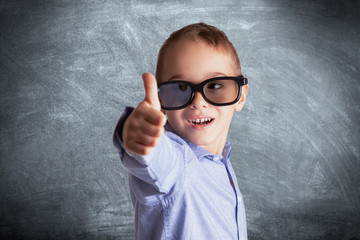 Young boy with eyeglasses in front of a school chalkboard showing thumbs up. Eye examination,...