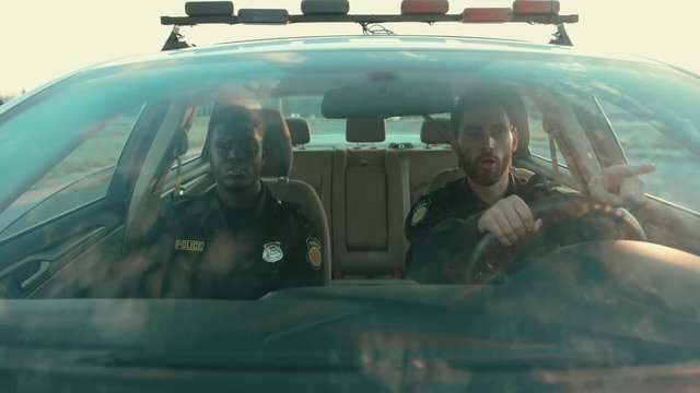 Curious policeman driving patrol car with his colleague to the emergency call at daytime. Front view afro-american cop talking walkie-talkie. Police on duty.
