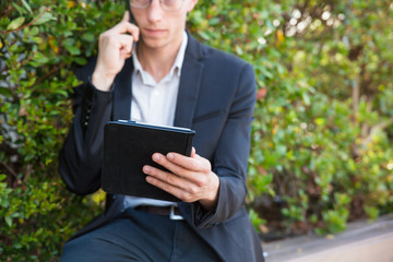 Serious professional using tablet and calling on phone. Young business man sitting outside, holding mobile computer and talking on cell. Working outdoors concept