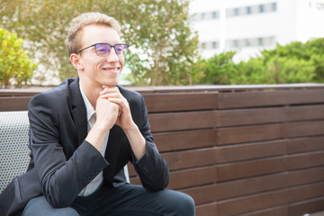 Fototapeta premium Cheerful manager enjoying work break outside. Happy young business man in eyeglasses and jacket sitting on bench, leaning chin on hands, looking away and smiling. Business portrait concept
