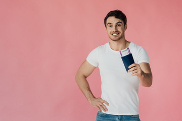 front view of smiling muscular man in white t-shirt holding passport and air ticket isolated on pink