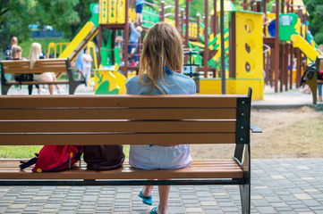 Young mother sits on a bench near the playground and looks after the child