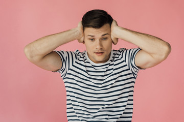 front view of young man in striped t-shirt covering ears isolated on pink
