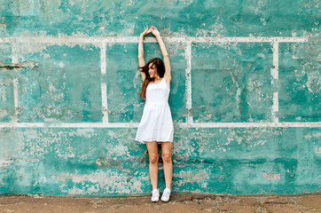 Fototapeta na wymiar Young brunette girl with long hair in white dresses is standing on the turquoise wall