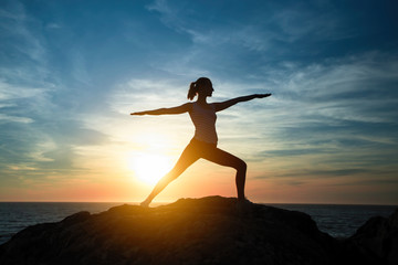 Yoga woman meditating in Assane the Warrior position. Silhouette on the sea coast during a beautiful sunset.