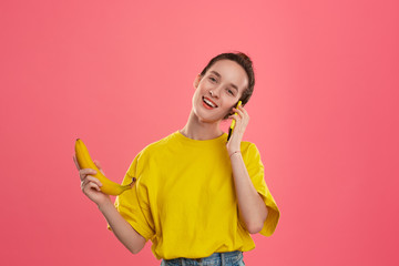Smiling model with a hair bun in a yellow t-shirt holding banana speaking on a yellow smartphone.