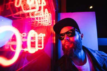 Hipster handsome man on the city streets being illuminated by neon signs. He is wearing leather...