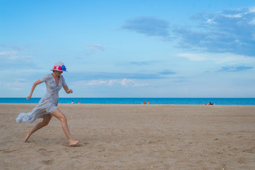 Fototapeta na wymiar A joyful middle-aged woman in a dress runs and jumps along the seashore of the ocean. Joy, pleasure, rest, freedom concept.