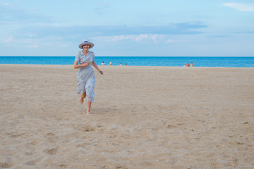 A joyful middle-aged woman in a dress runs and jumps along the seashore of the ocean. Joy, pleasure, rest, freedom concept.