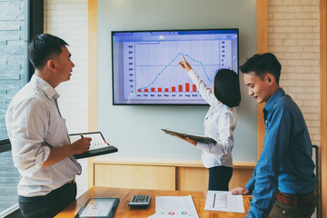 Beautiful businesswoman gives report or presentation to her business colleagues in the conference room. She show graphic charts and company's growth on the wall TV.