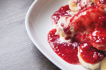 Closeup Vanilla ice cream poured with strawberry or raspberry jam with sliced banana slices on a white plate on a dark wooden table. You can do this at home on vacation