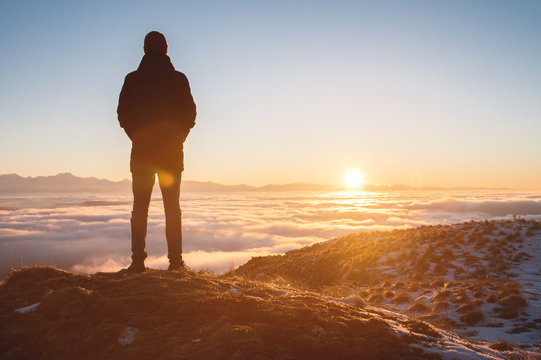 View From the back. A lonely standing man high in the mountains looks at the setting sun and the sunset horizon with a valley filled with clouds. The concept of tourism travel and male loneliness - Powered by Adobe