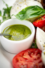 Basil pesto sauce in a small bowl, with fresh basil leaves, tomato and mozzarella cheese, close up