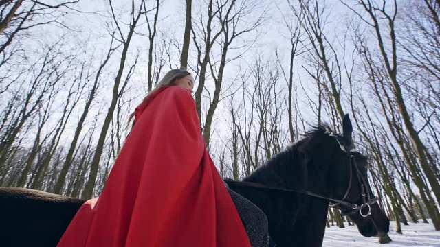 From below attractive horsewoman with long hair wearing red cloak riding strong black horse in winter forest and periodically looking at camera and smiling