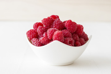 The porcelain bowl with fresh raspberry fruit on the white wooden table.