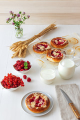 White table with yeasty sweet pastry with fresh currants and raspberries and streusel topping, glass and pitcher of milk, sheaf of wheat, small glass vase with flowers. Summer fresh fruit appetizer.
