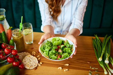 Young happy woman eating salad in the beautiful interior with green flowers on the background and fresh ingredients on the table. Healthy food concept