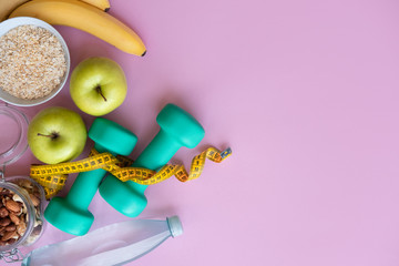 Fitness concept with dumbbells, nuts, bananas, apples and skipping rope on pink background with copy space