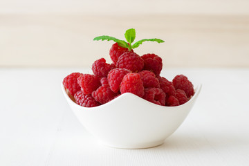 The porcelain bowl with fresh raspberry fruit embellished with fresh green mint on the white wooden...