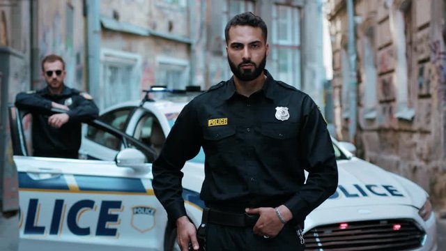 Face young man cops stand near patrol car look at camera serious enforcement officer police uniform auto safety security communication control policeman close up slow motion