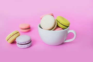 Creative image of sweet colorful French macaroon biscuits in a white mug on pink background.