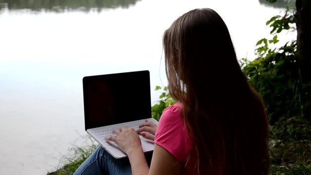Cute Blonde girl sitting on the lake and working with laptop in summer or sping autumn day against beautiful landscape of northern lake and forest. Freelance, work and travel concept. call the phone