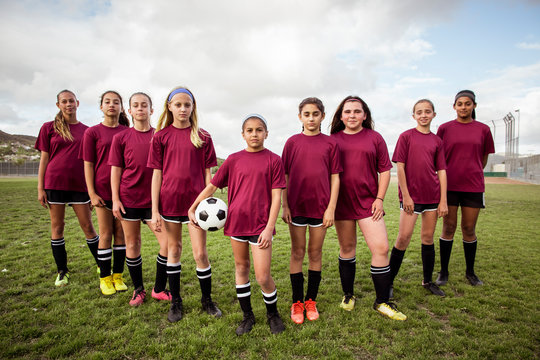 Full length portrait of confident girls in soccer uniform standing on field during sports competition against sky