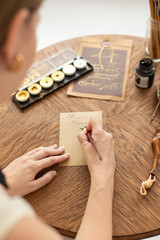 Attractive woman calligrapher is holding a pen with ink and signs a card in handwritten font....
