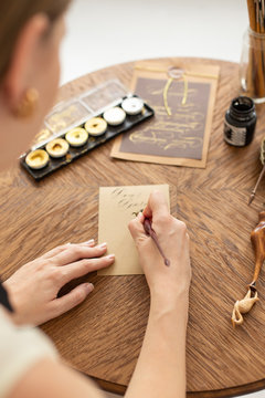 Attractive woman calligrapher is holding a pen with ink and signs a card in handwritten font. Close-up. Soft focus.