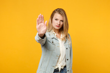 Portrait of serious young woman in denim casual clothes looking camera showing stop gesture with palm isolated on yellow orange wall background in studio. People lifestyle concept. Mock up copy space.