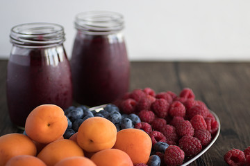 Fruit and berry smoothies and fresh apricots, raspberries and blueberries on an oval metal plate. Gray background.