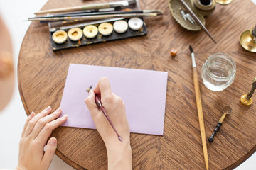 Woman calligrapher is holding a pen with ink and signs a letter in handwritten font. Close-up. Soft focus. Top view.