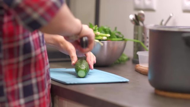Young woman housewife cuts cucumber on a blue plastic cutting board. Preparing food ingredients on a stone countertop at a home kitchen. 4k 50 frames per second narrow depth of field close up.