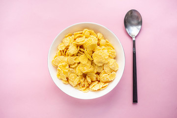 Cornflakes dry breakfast in a white bowl with a spoon on a pink backgroud. top view