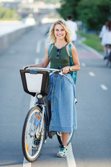 Photo of young blonde in long denim skirt standing next to bike on road in city