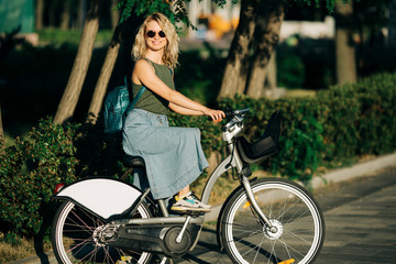 Fototapeta na wymiar Image of young blonde in sunglasses and long denim skirt standing on bike next to green bushes in city