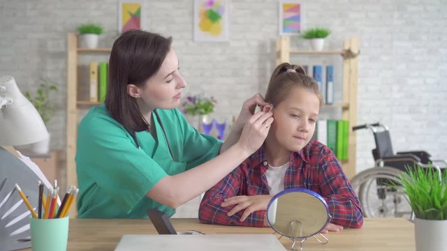 doctor otolaryngologist helps to put on the hearing aid to a hearing impaired teenage girl