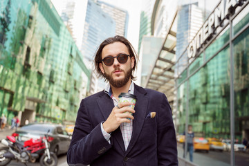 Photo of brunet with beard and earring in ear from glass in hand in city against background of buildings, cars