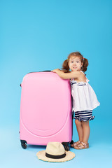 Happy child girl tourist packs clothes into a suitcase for travel, summer vacation. Toddler kid in straw hat  holding a pink suitcase on a blue background