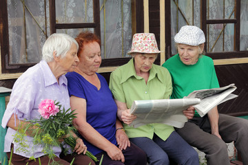 Four Russian women sit on a bench at the village house and actively discuss the news from the newspaper. Country life.