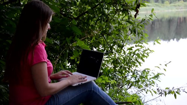Cute Blonde girl sitting on the lake and working with laptop in summer or sping autumn day against beautiful landscape of northern lake and forest. Freelance, work and travel concept. call the phone