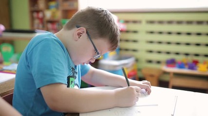 The boy with glasses writes in pencil in a notebook. Pupil in elementary school solves a logical problem. A lesson in elementary school on logic and mathematics. Preschool education.